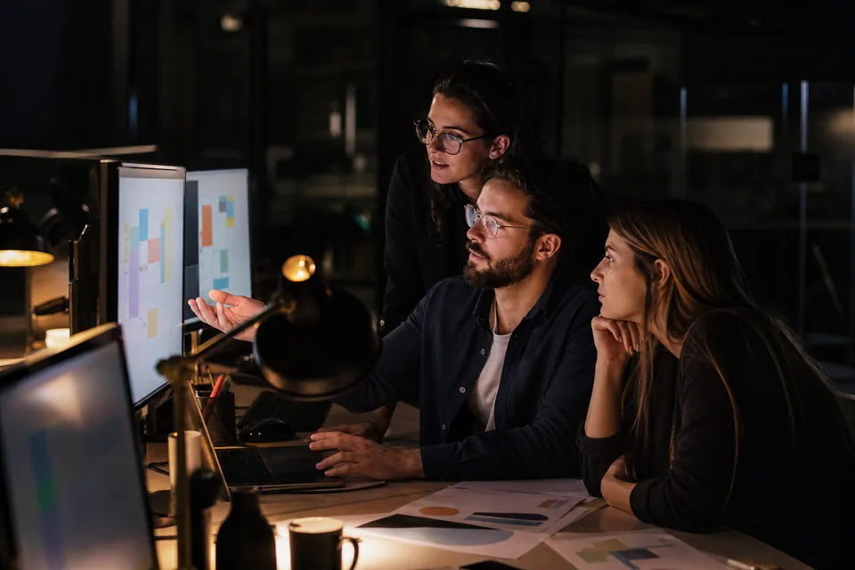 Three marketers working at night on monitors with content calendars under brass desk lamps